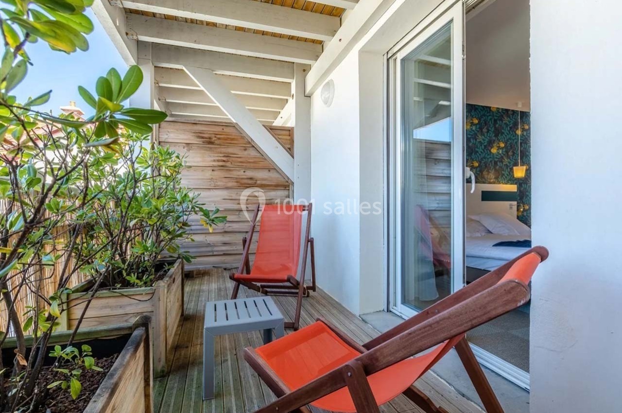 Balcon en bois avec deux chaises orange, une table basse grise et des plantes en pot, donnant sur une chambre lumineuse.