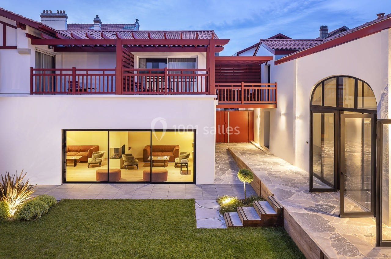 Façade d'une maison moderne avec grandes baies vitrées, terrasse en pierre et jardin éclairé au crépuscule.
