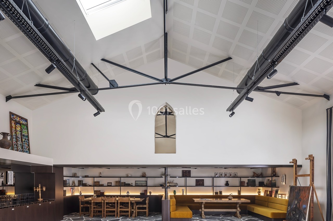 Salle lumineuse avec plafond voûté, grandes poutres noires, tables en bois, banquettes jaunes et sol en pierre.