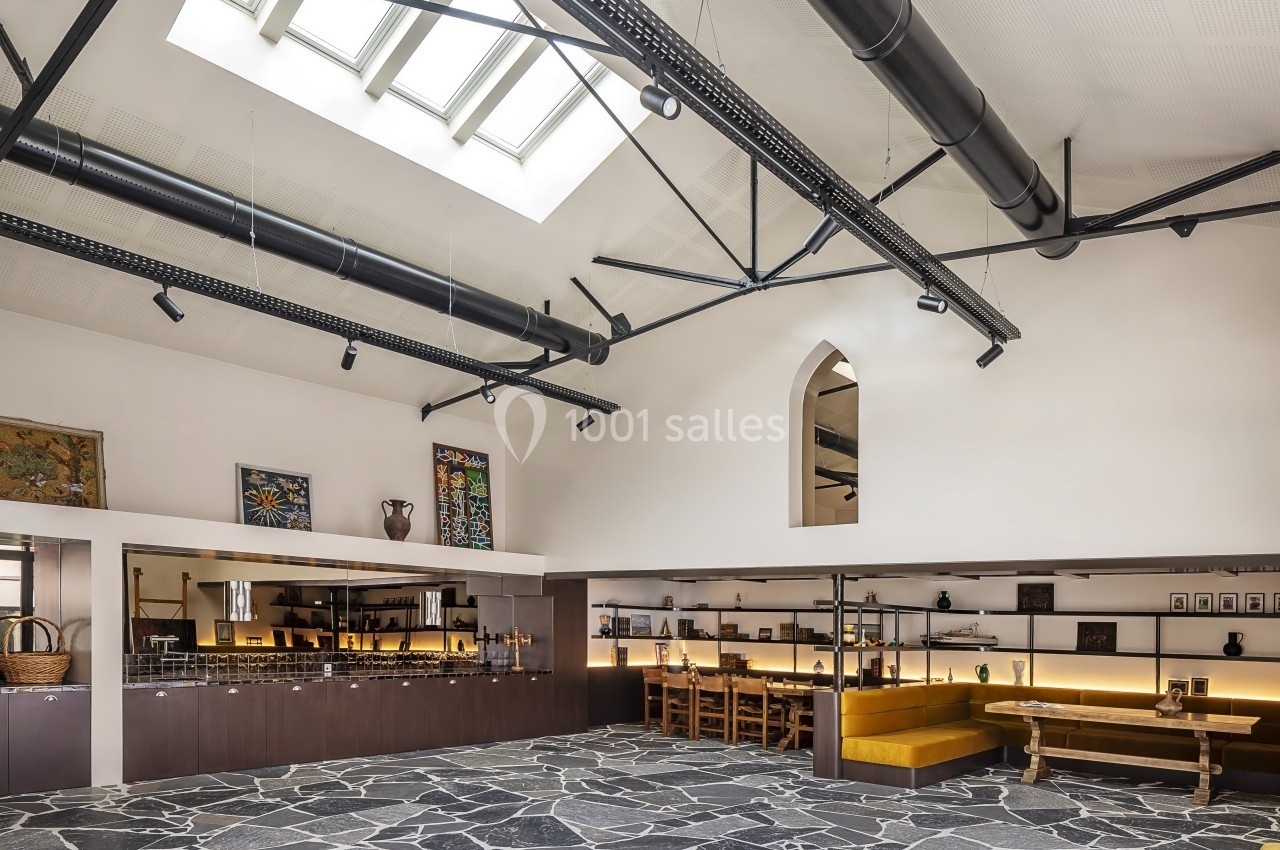 Salle lumineuse avec sol en pierre, plafond haut avec poutres apparentes, tables en bois et étagères décoratives.