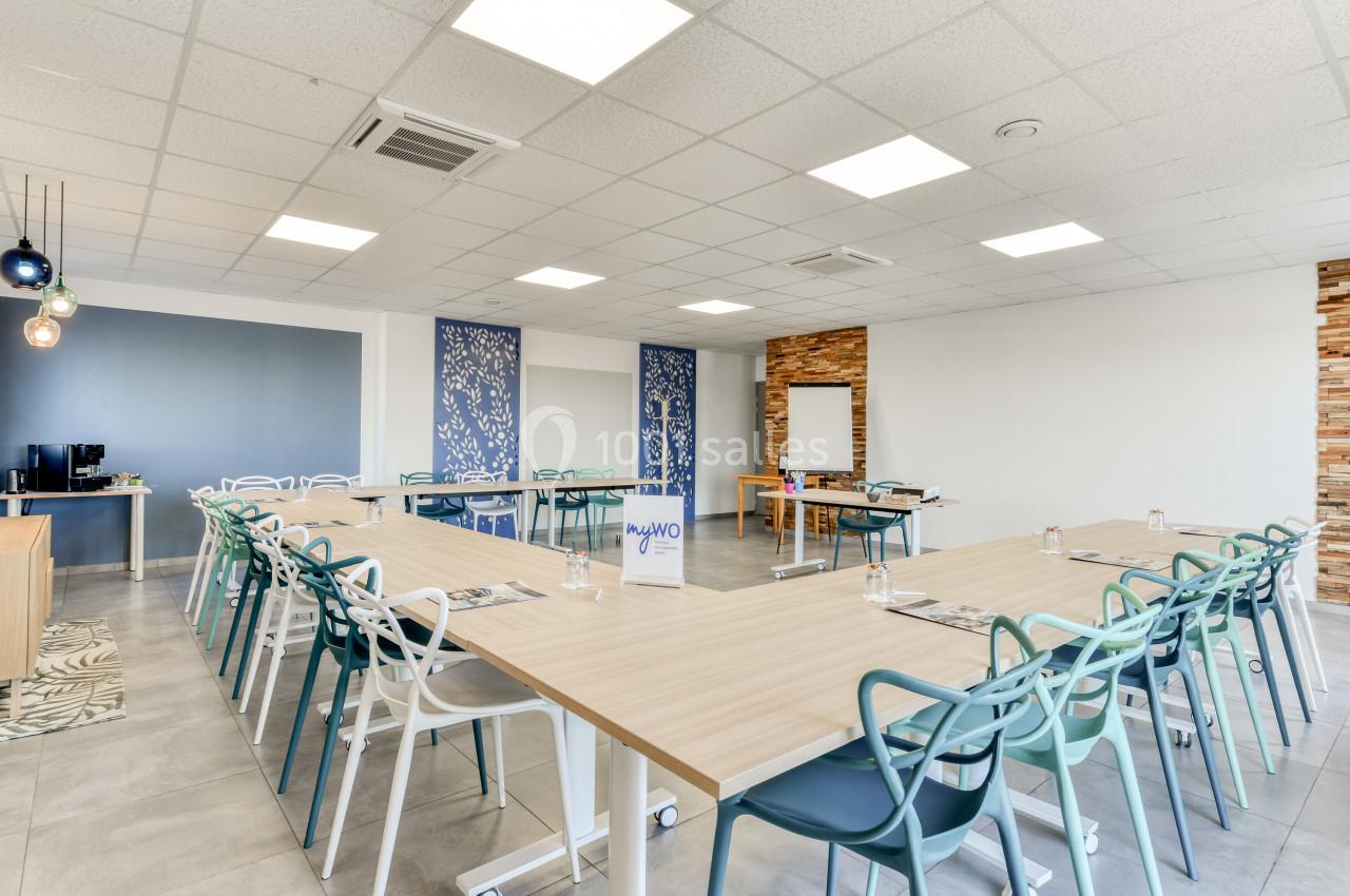 Salle de réunion lumineuse avec tables disposées en U, chaises colorées et tableau blanc au fond.