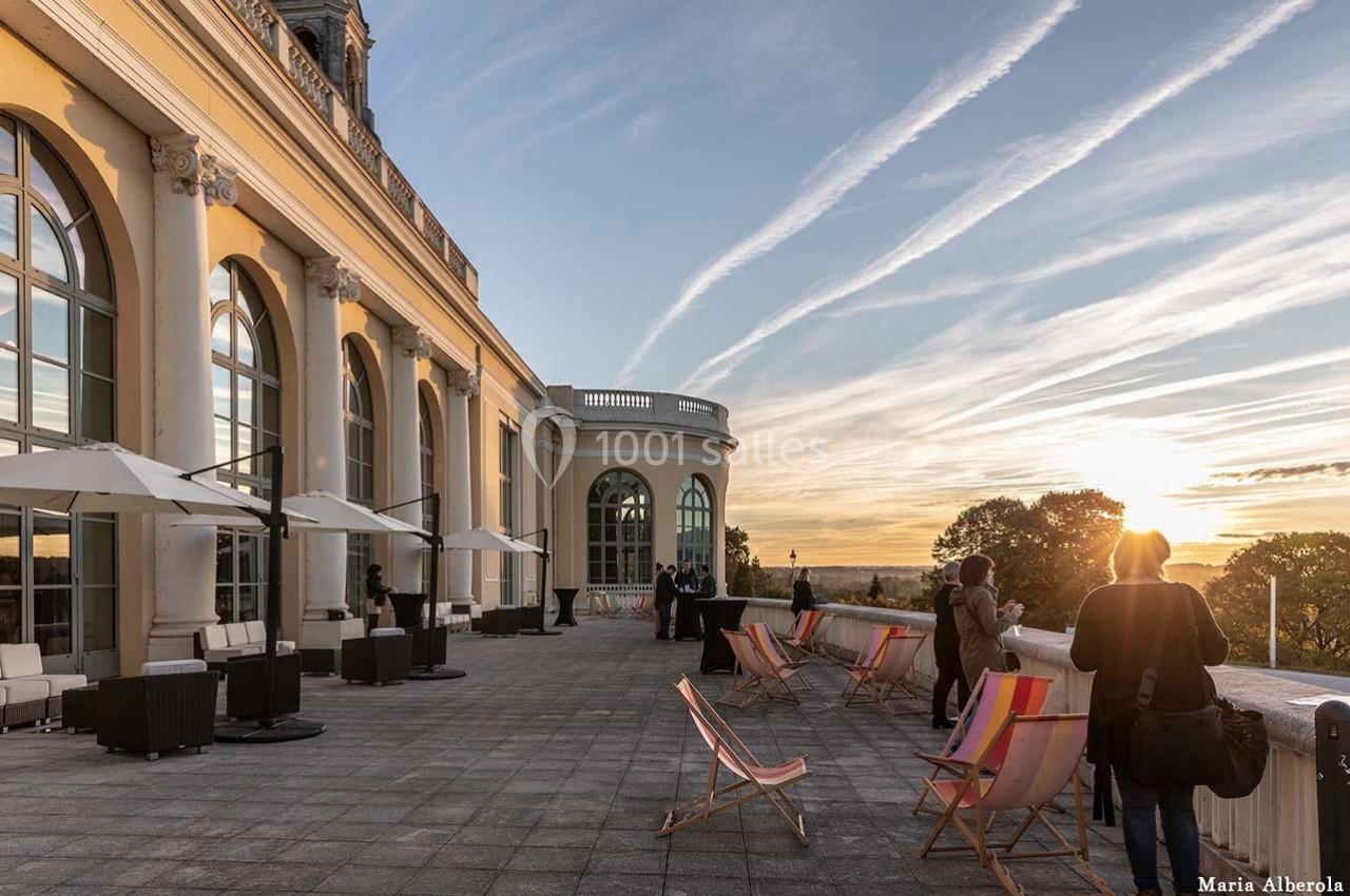 Terrasse avec chaises longues, parasols et vue sur un coucher de soleil, près d'un bâtiment aux grandes baies vitrées.