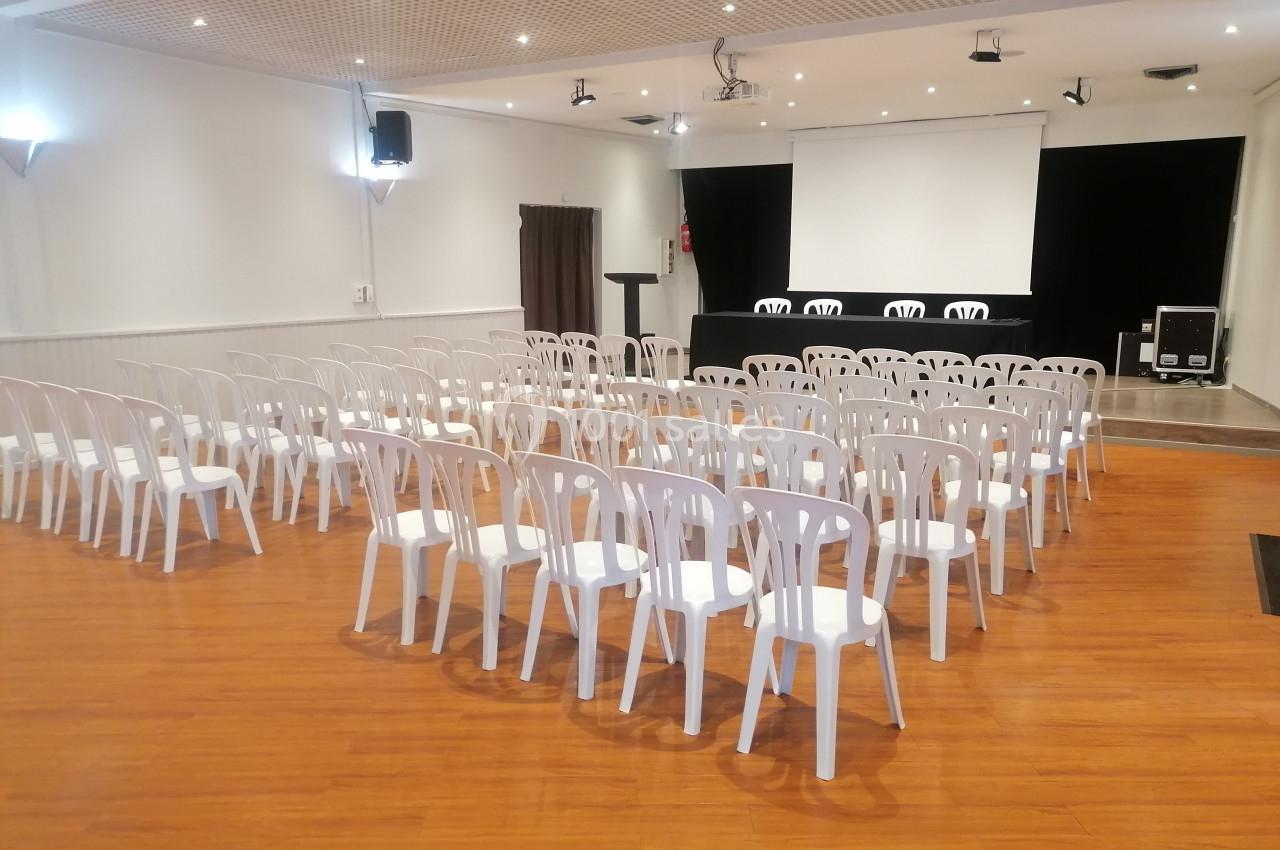 Salle de conférence avec des rangées de chaises blanches en plastique, un podium et un écran de projection à l'arrière.