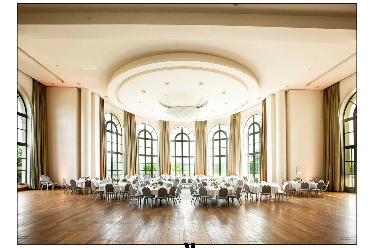 Salle de réception lumineuse avec parquet en bois, grandes fenêtres cintrées et tables rondes dressées.