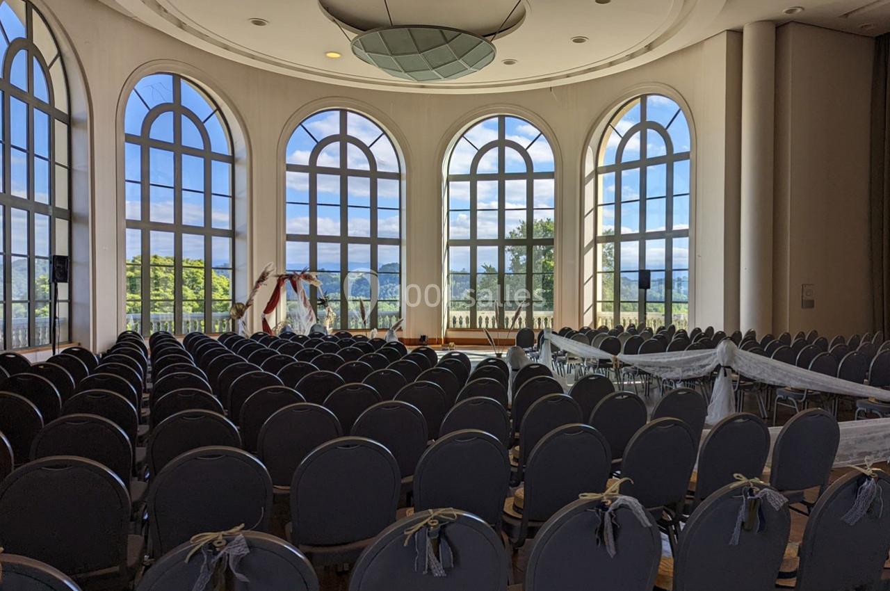 Salle de réception lumineuse avec de grandes fenêtres cintrées, rangées de chaises et une arche décorative au fond.