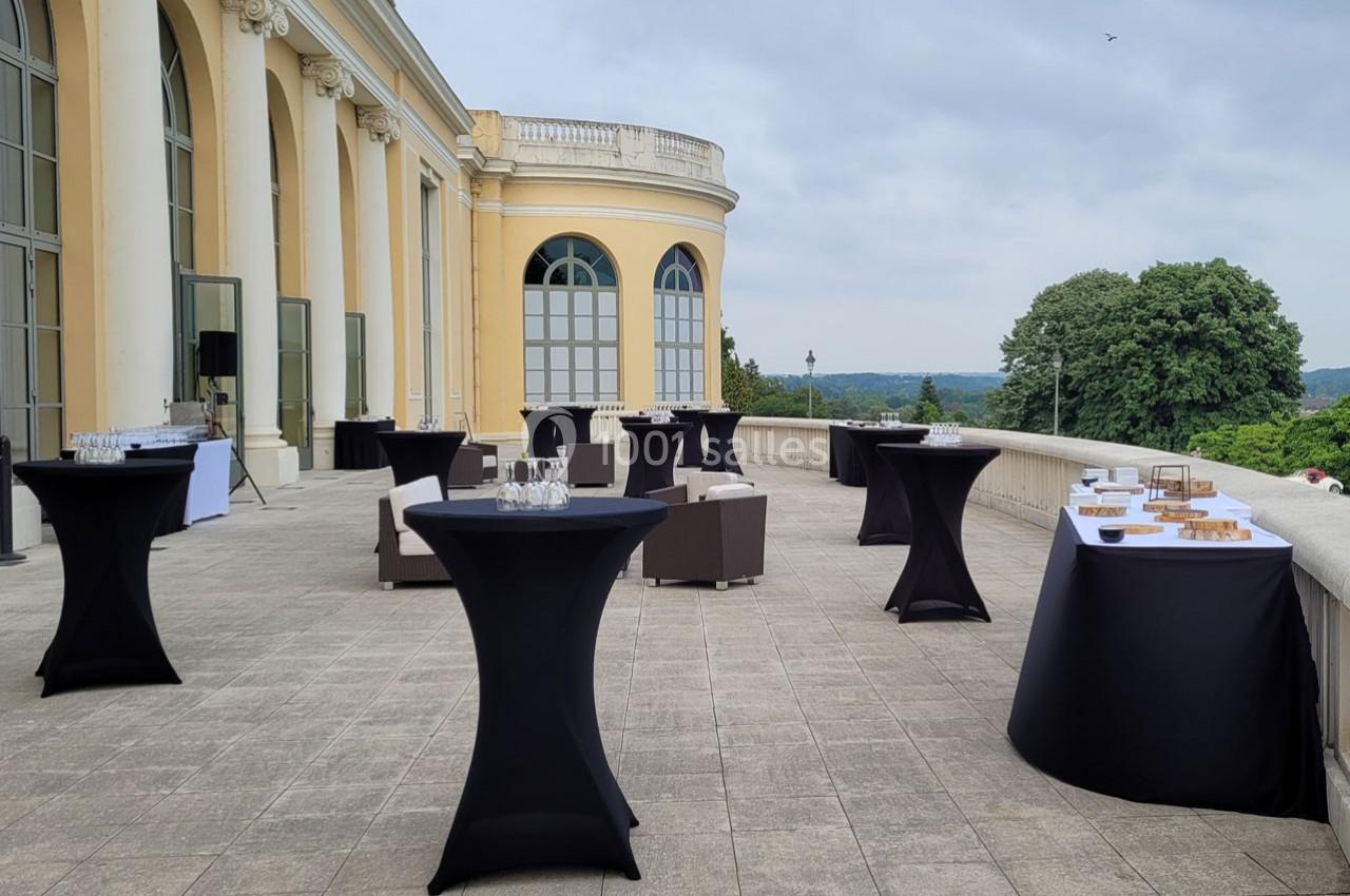 Terrasse avec tables hautes noires, canapés et buffets dressés, donnant sur un paysage verdoyant sous un ciel nuageux.