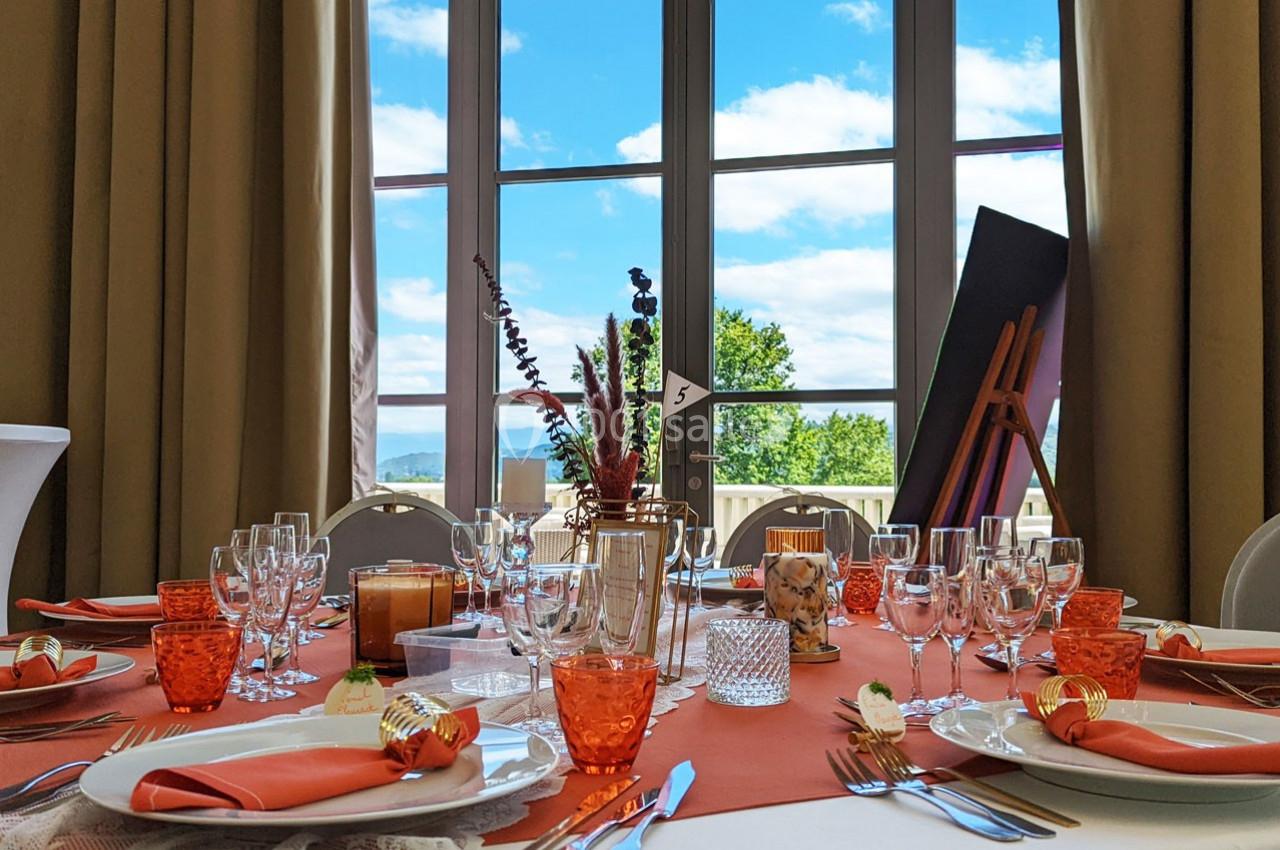 Table dressée avec vaisselle élégante et décorations orange, devant une grande baie vitrée offrant une vue sur un paysage.