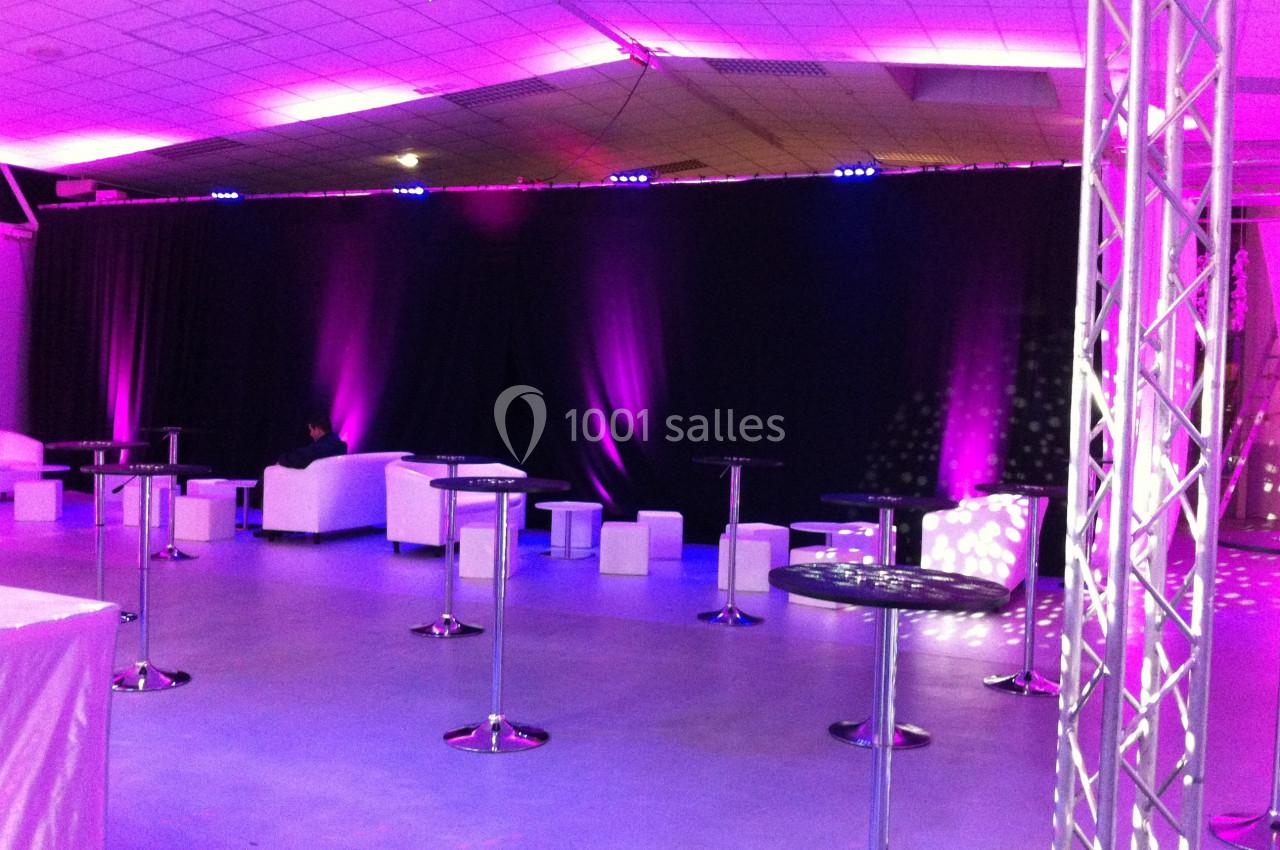 Salle de réception éclairée par des lumières violettes, avec tables hautes, tabourets et canapés blancs.