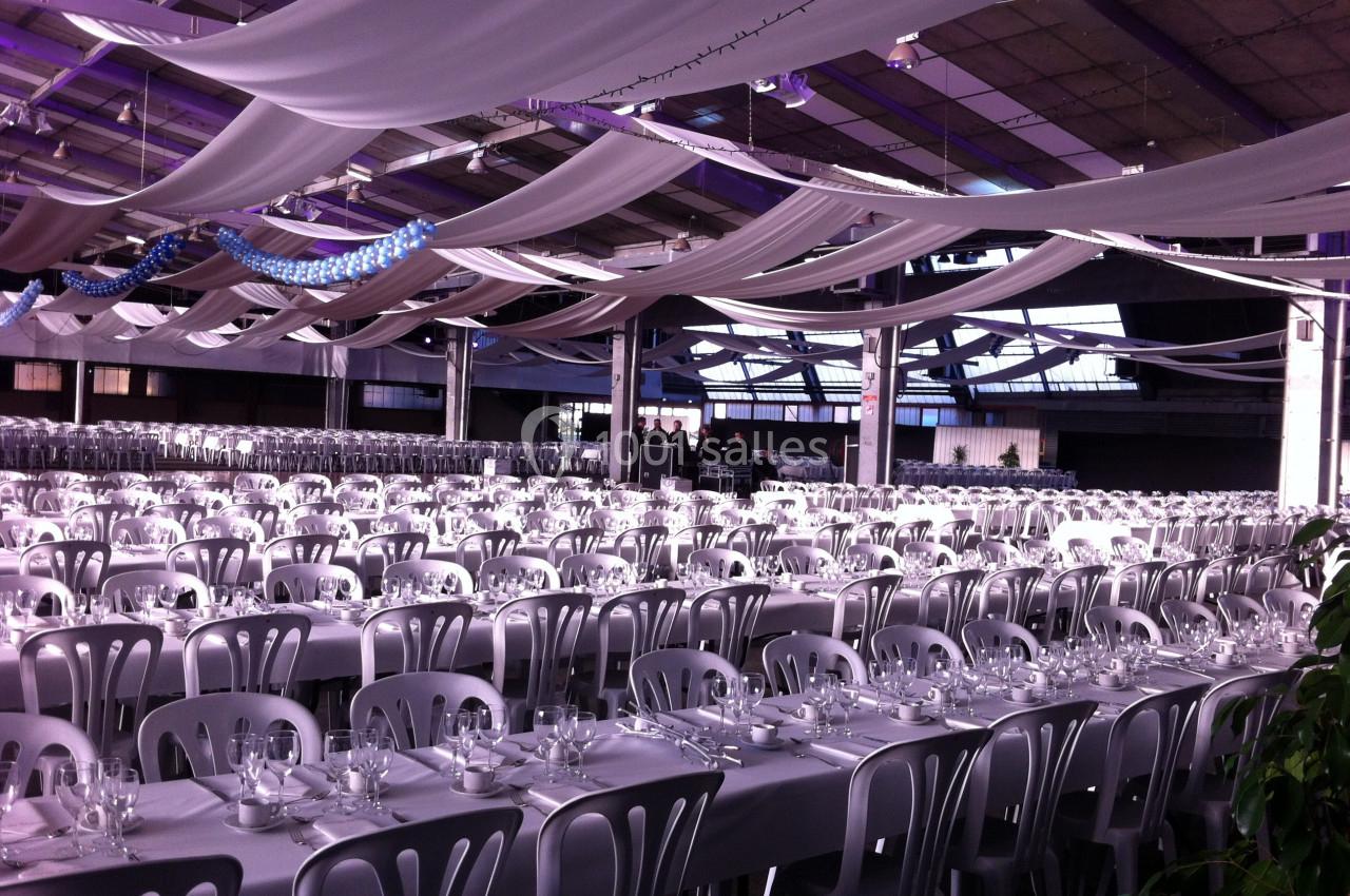 Grande salle décorée avec des drapés suspendus, aménagée avec de longues tables dressées et alignées pour un événement.