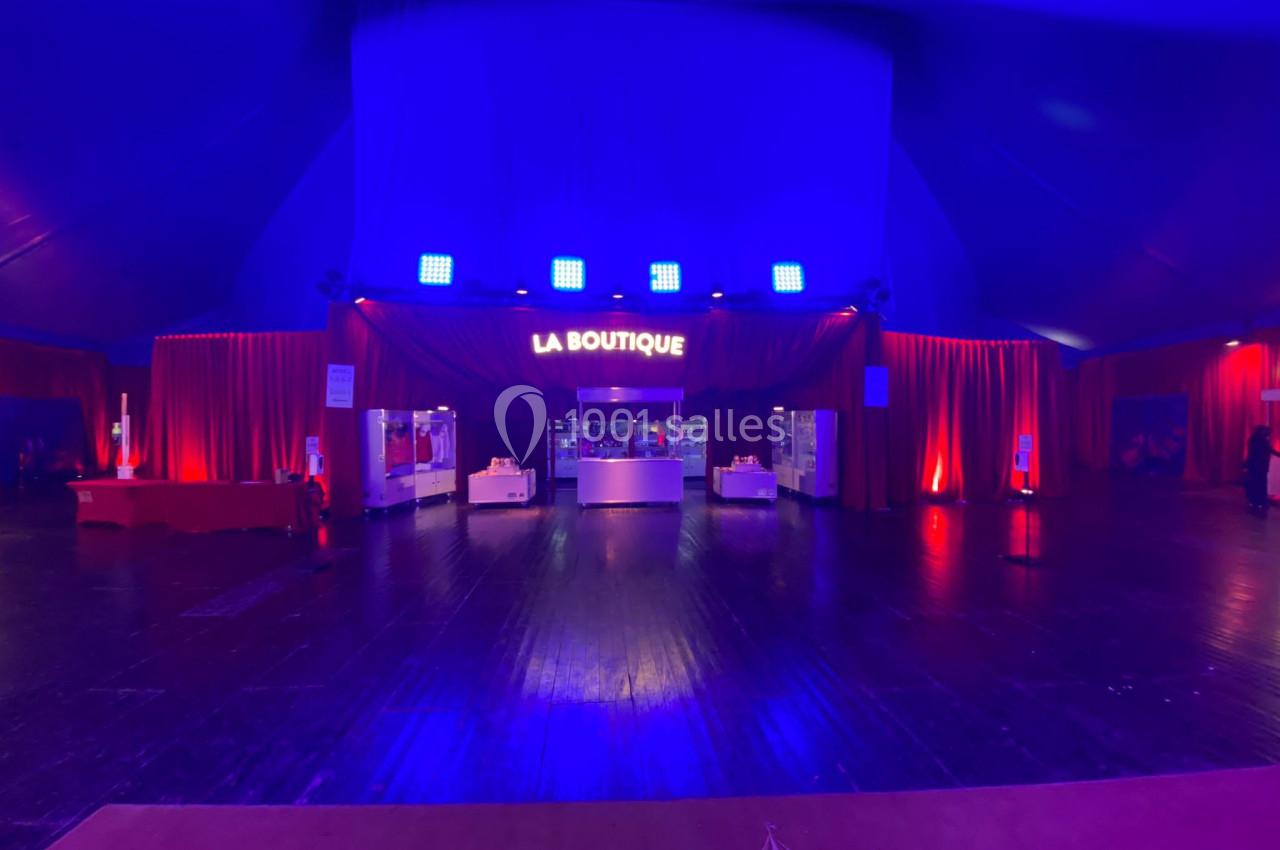 Intérieur d'un chapiteau avec un stand lumineux ’La Boutique’ entouré de rideaux rouges et éclairages bleus.