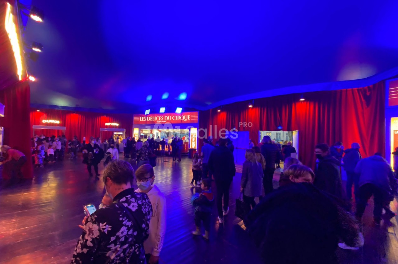 Foule rassemblée dans un chapiteau éclairé de lumières bleues et rouges, avec des stands visibles en arrière-plan.