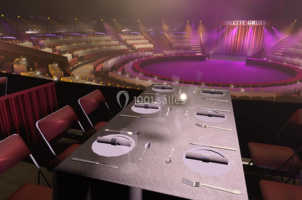Salle de spectacle circulaire avec scène centrale, tables dressées au premier plan et gradins éclairés en arrière-plan.