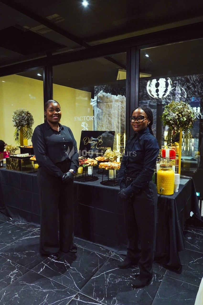 Deux personnes en tenue noire devant une table garnie de boissons, plats et décorations florales dans un espace intérieur.