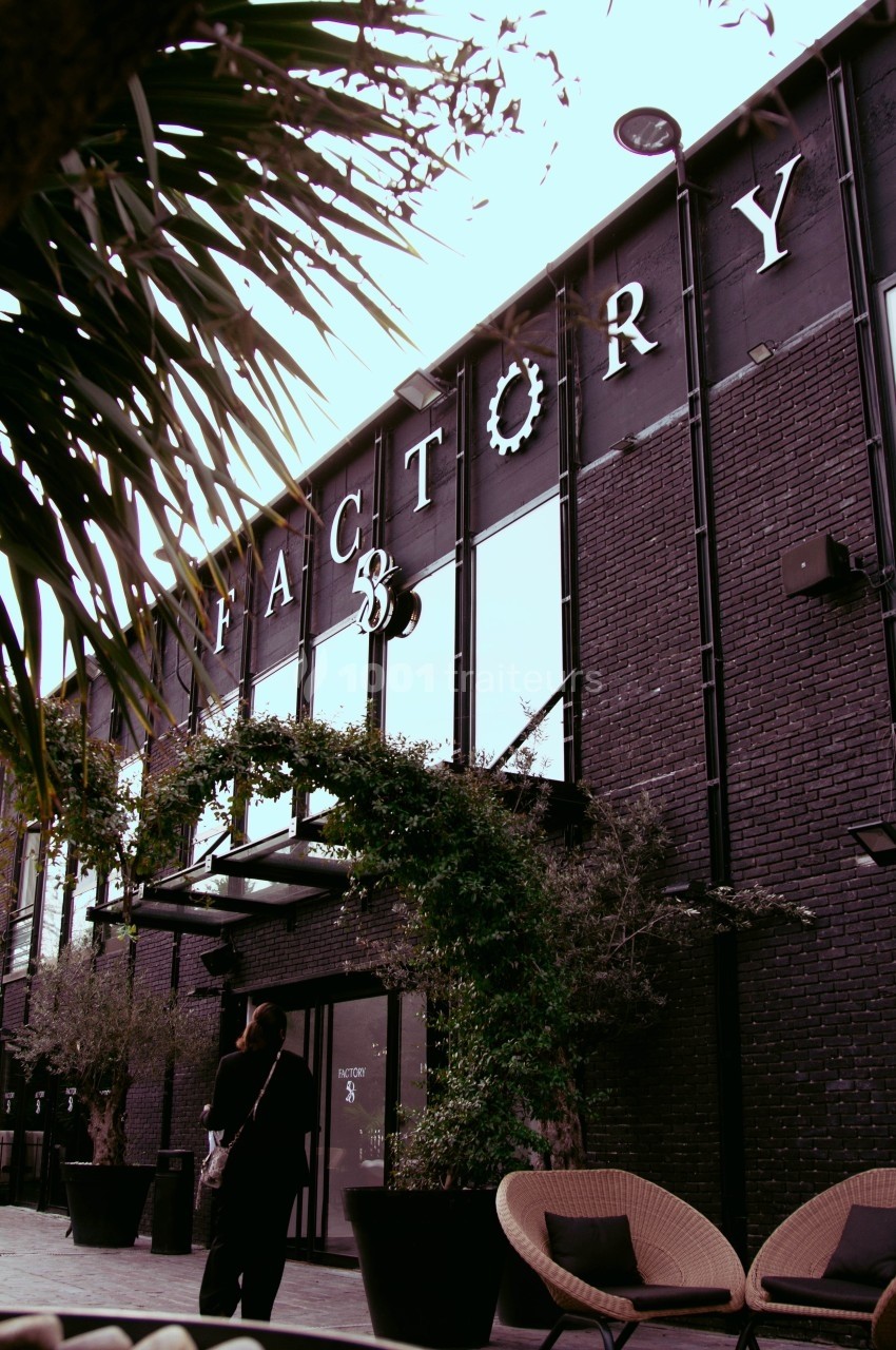 Façade d'un bâtiment en briques noires avec l'inscription ’Factory’, entouré de végétation et de mobilier extérieur.