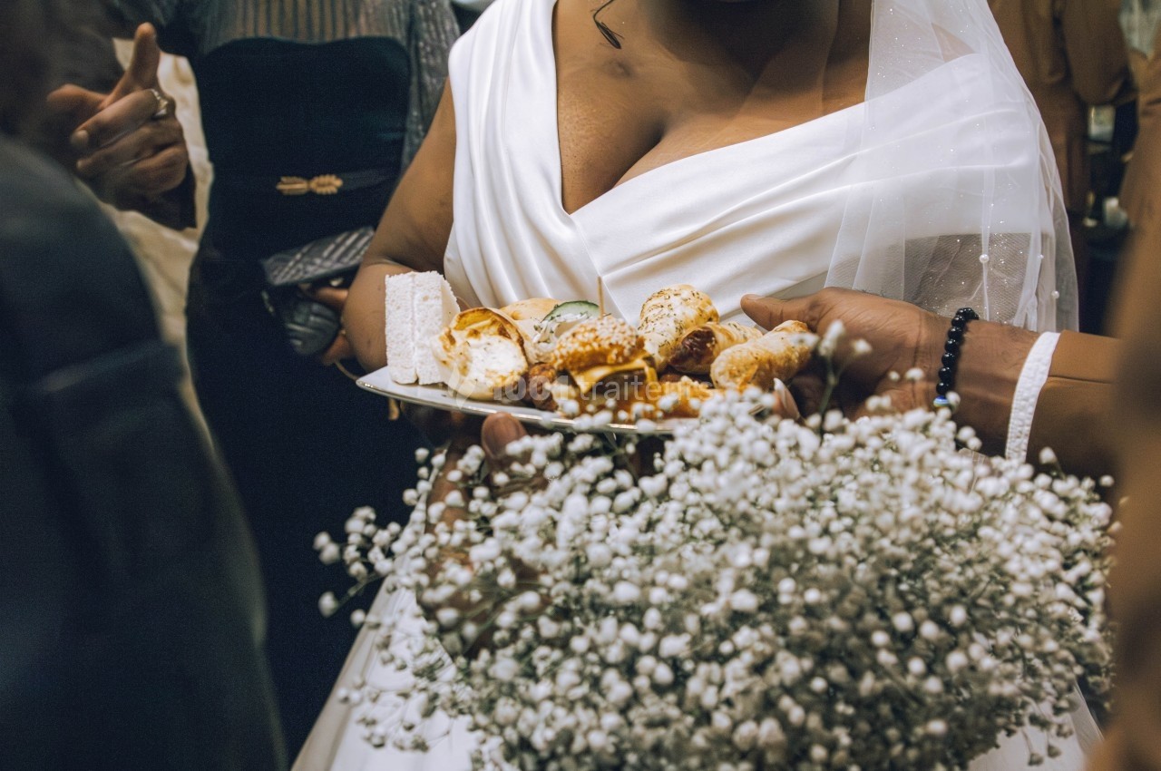 Une mariée en robe blanche tient un plateau de petits amuse-bouches, entourée de personnes et d'un bouquet de gypsophiles.