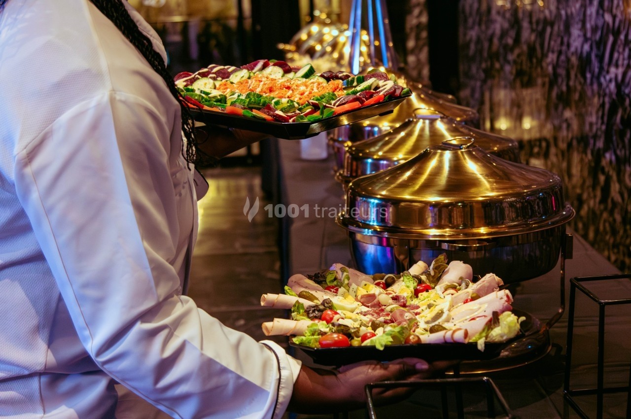 Un serveur porte des plateaux de charcuterie et de légumes devant un buffet avec des plats en métal doré.