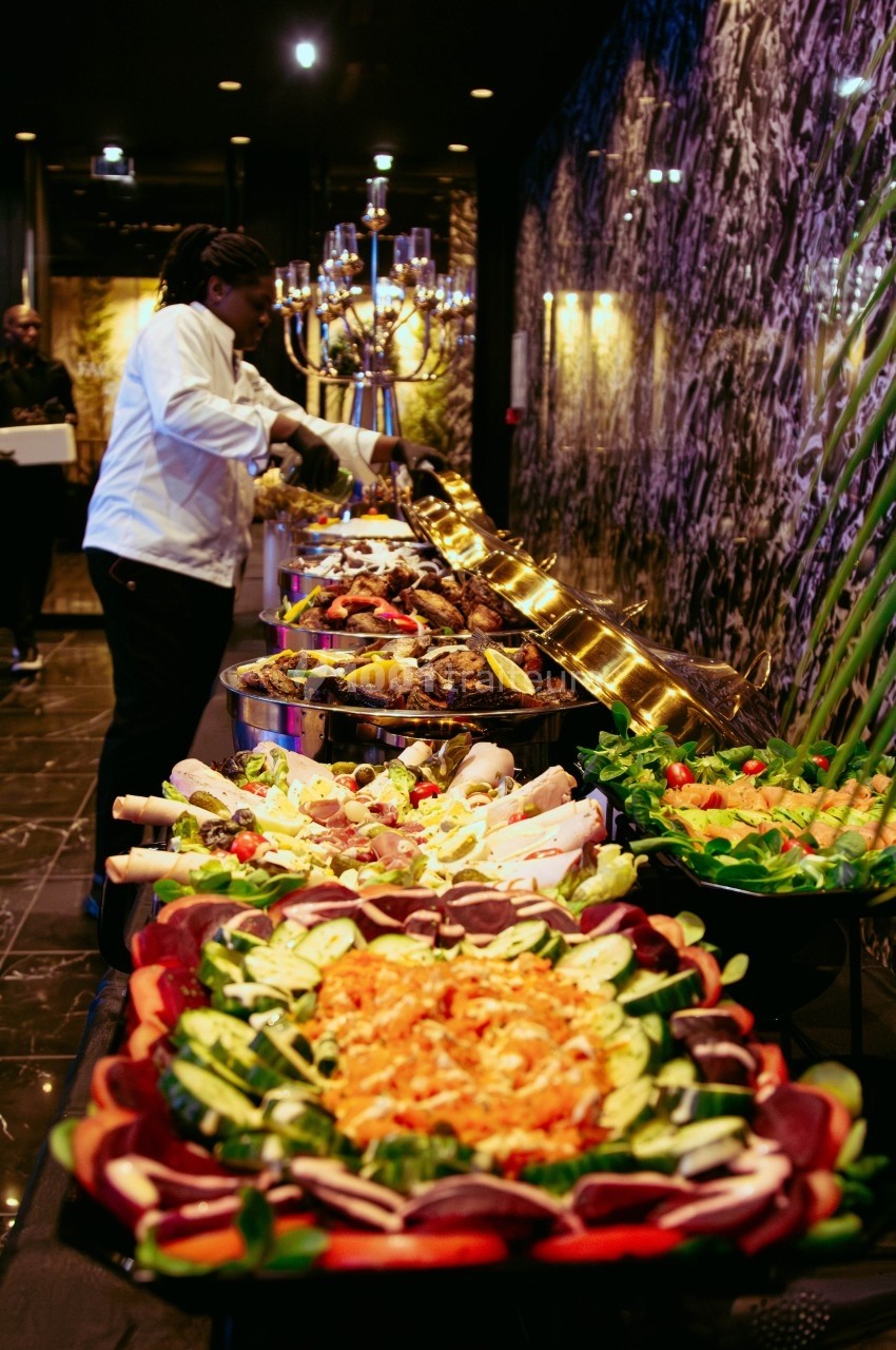 Buffet varié avec plats colorés, légumes frais, viandes grillées et chef en train de préparer les assiettes.