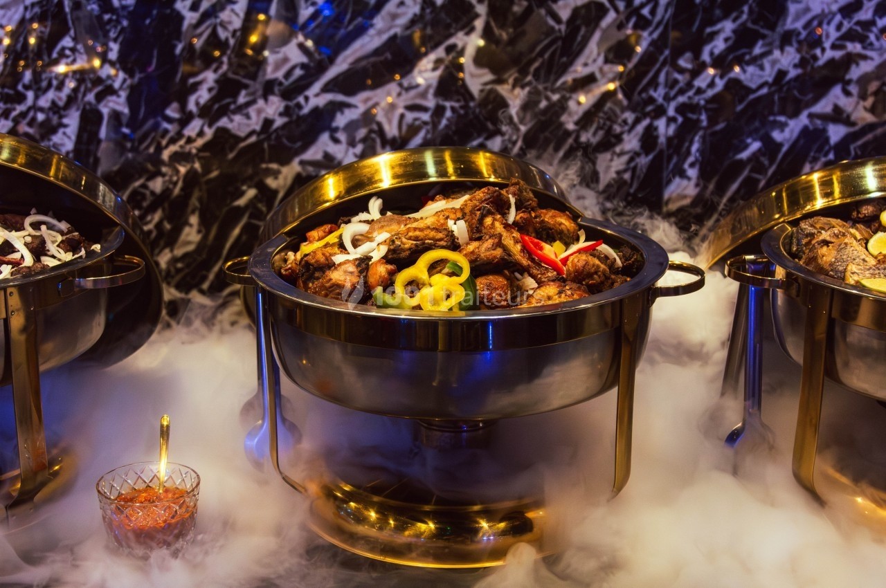 Buffet avec un plat chaud de viande garnie de légumes, présenté dans un récipient en métal sur un fond de fumée.