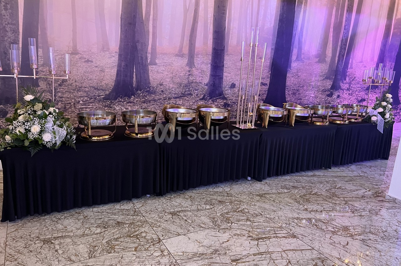 Table décorée avec des plats en métal doré, chandeliers et arrangements floraux, sur fond de forêt illuminée.
