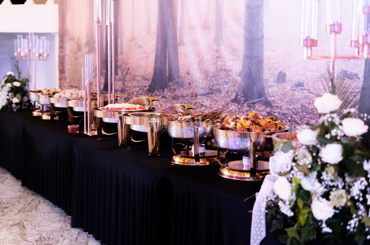 Buffet varié avec plats chauds et froids disposés sur une table noire, décorée de fleurs blanches et chandeliers.