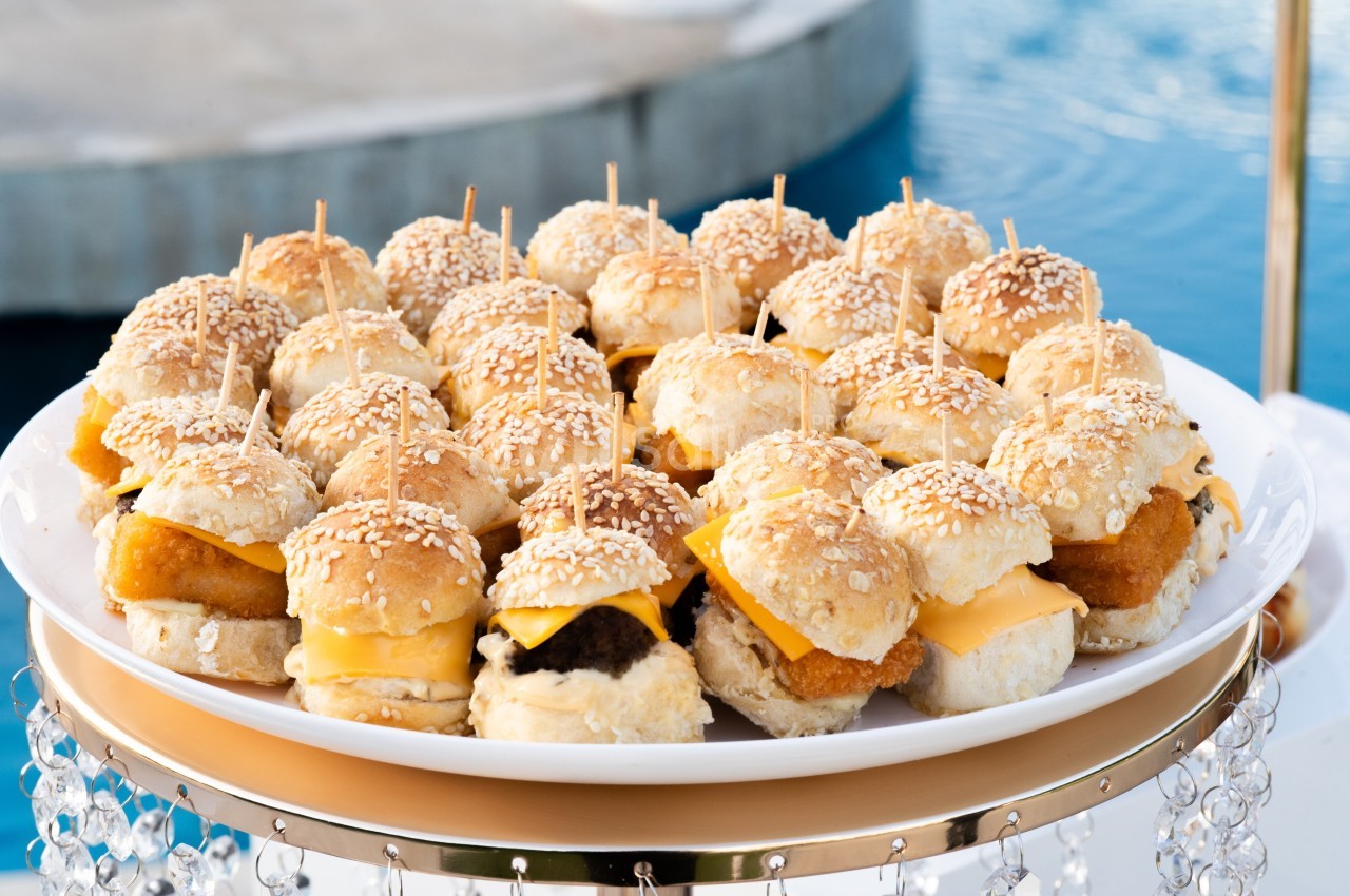 Plateau de mini-burgers variés avec fromage et pain aux graines de sésame, présenté près d'une piscine.