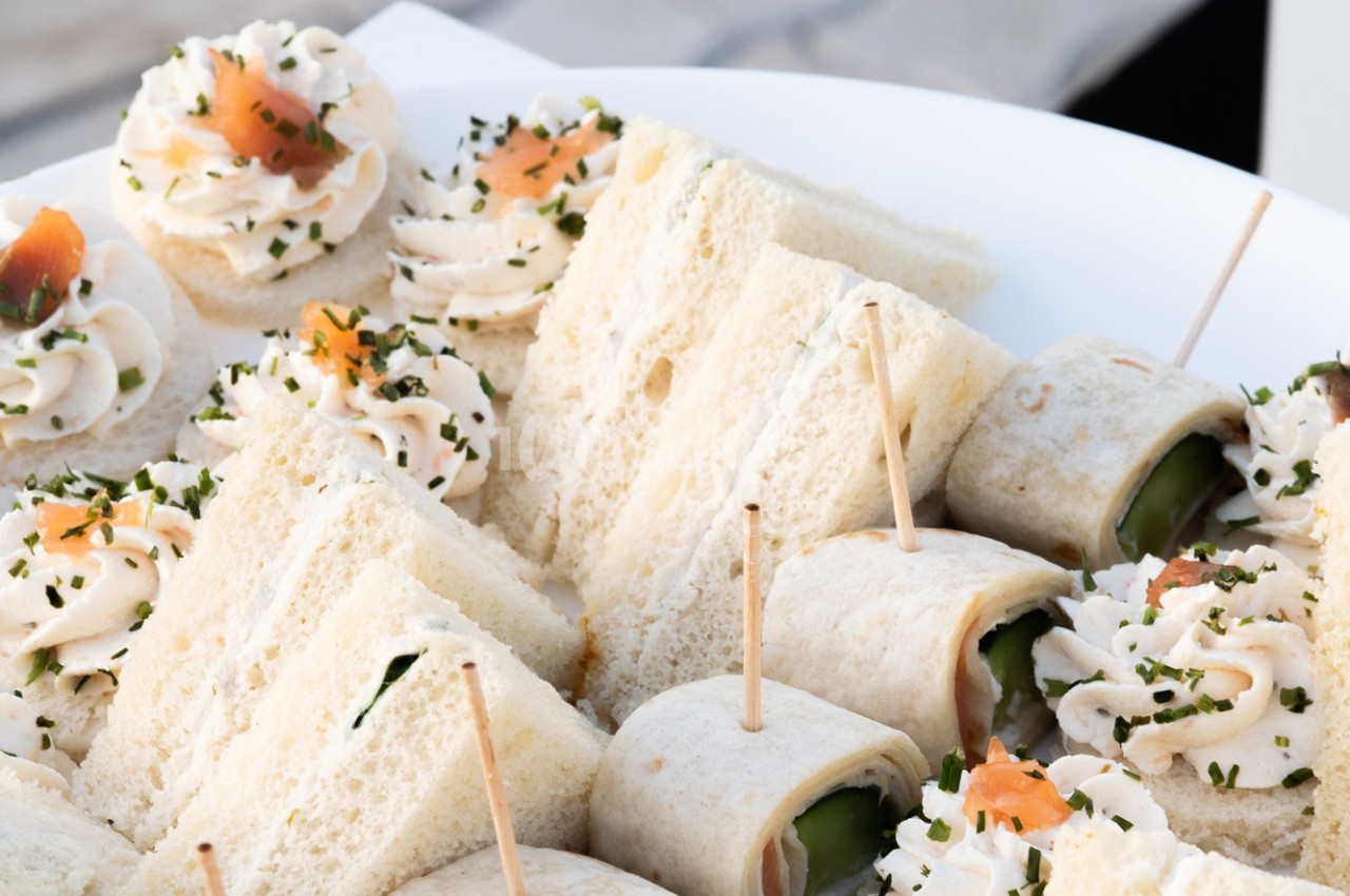 Assortiment de sandwichs, roulés au concombre et bouchées au saumon sur un plateau blanc.