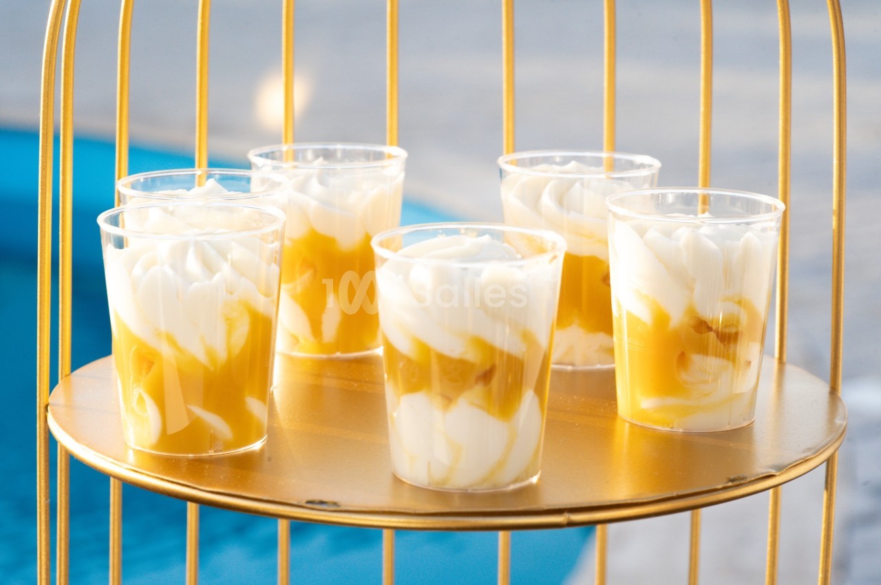 Cinq verrines de dessert à base de crème et coulis jaune, posées sur un plateau doré près d'une piscine.