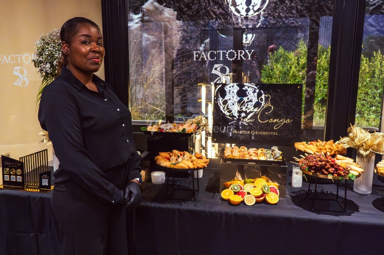 Une femme en tenue noire se tient devant un buffet garni de plats variés et décorés, dans un cadre élégant.