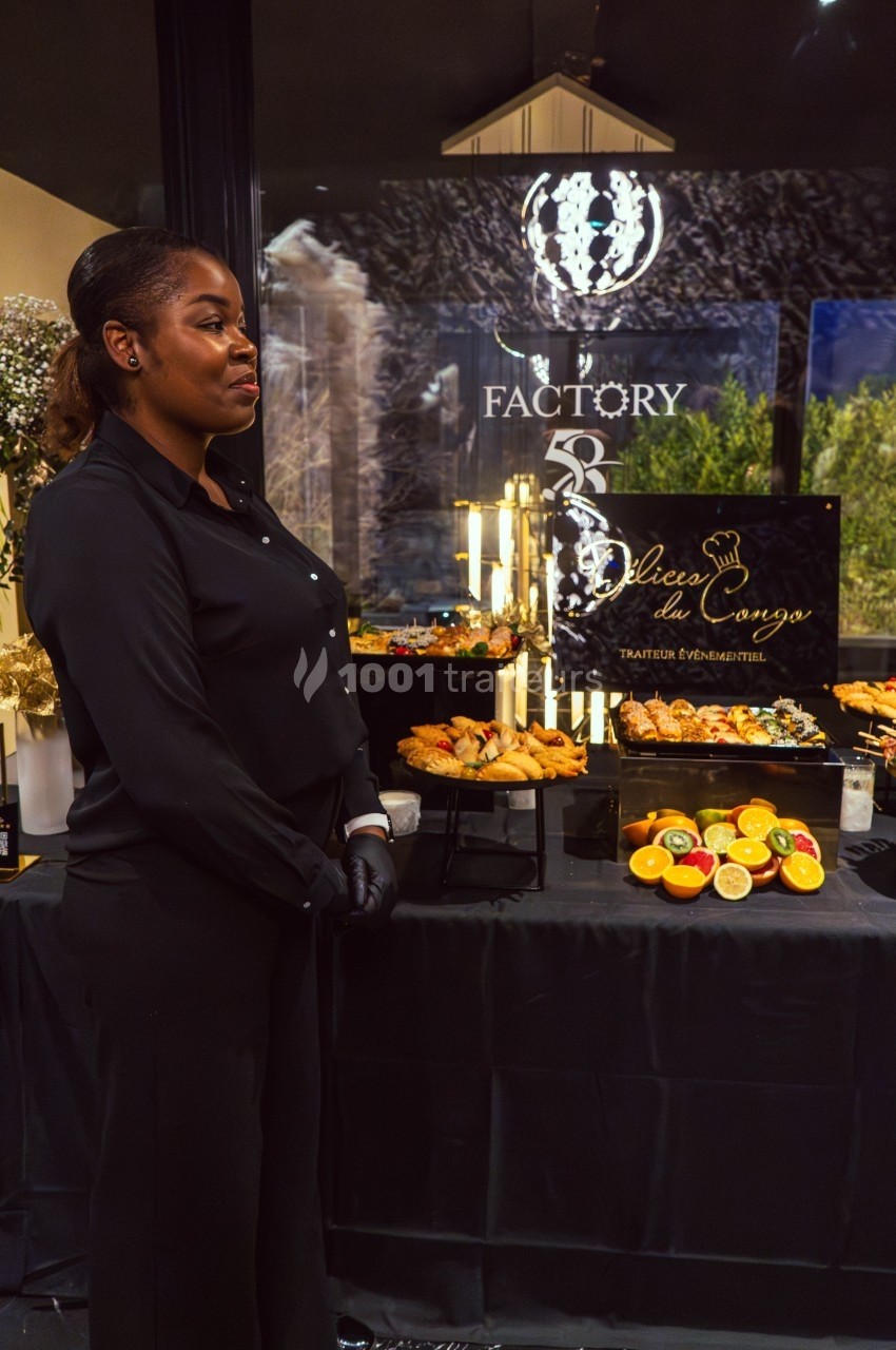 Une femme en tenue noire se tient devant une table de buffet garnie de fruits et de plats variés.