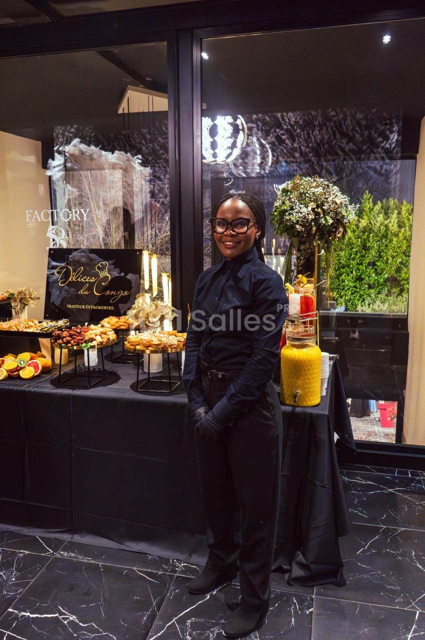Une personne souriante en tenue professionnelle devant une table garnie de desserts et de boissons dans un cadre élégant.