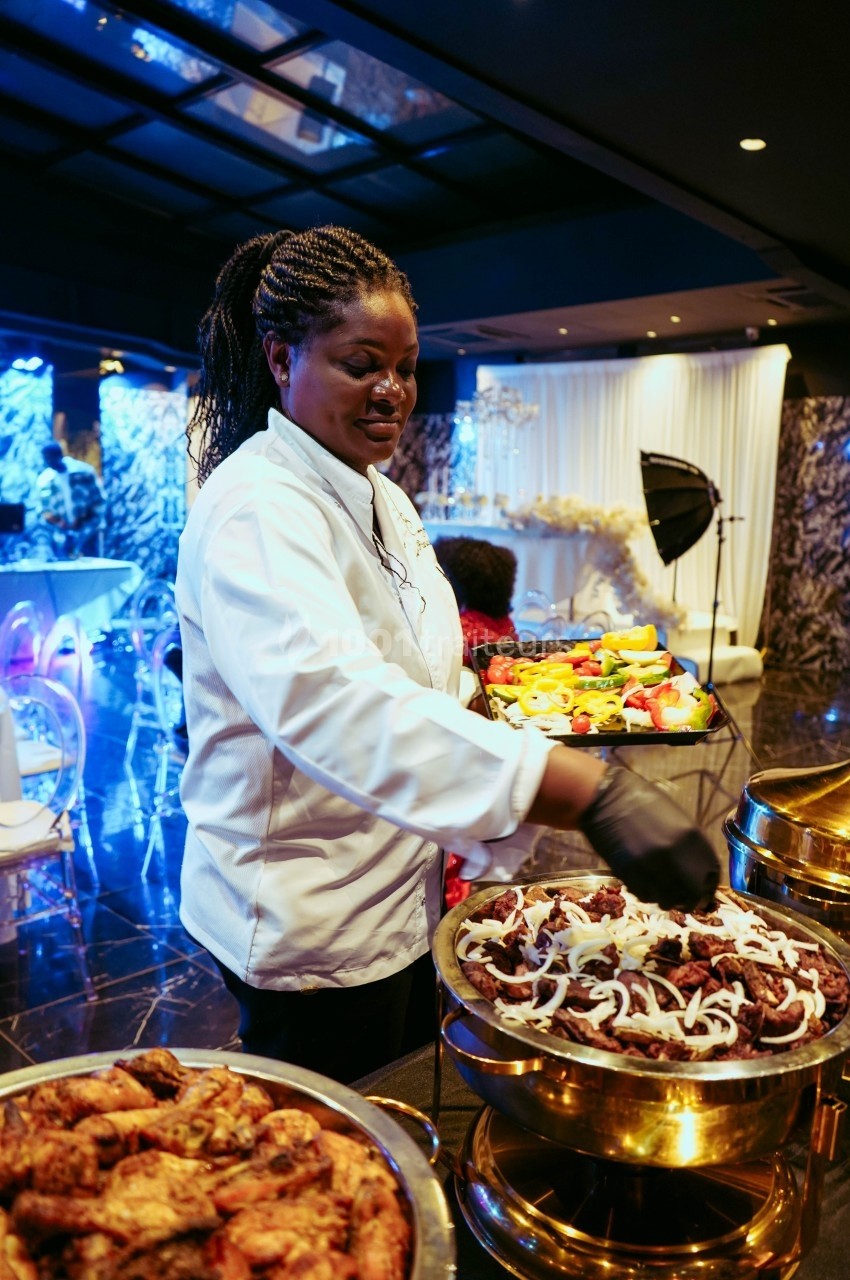 Une cheffe en tenue blanche dispose des plats garnis de viande et légumes dans une salle de réception élégante.