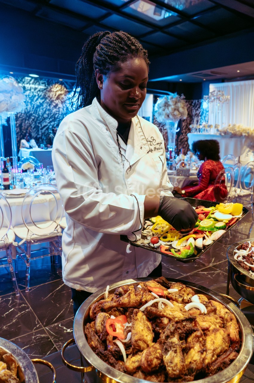 Une cheffe en tenue blanche dispose des légumes frais sur un plateau lors d'un buffet dans une salle élégante.