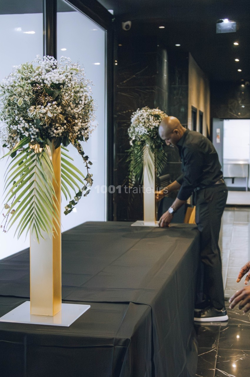 Un homme installe une table recouverte d'une nappe noire, décorée de grands bouquets sur des supports dorés.