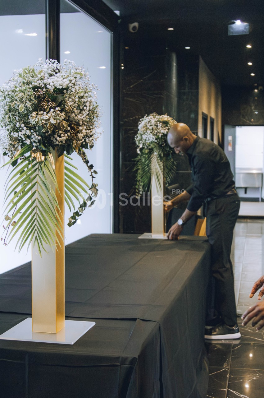 Un homme installe une table recouverte d'une nappe noire, décorée de grands bouquets sur des supports dorés.