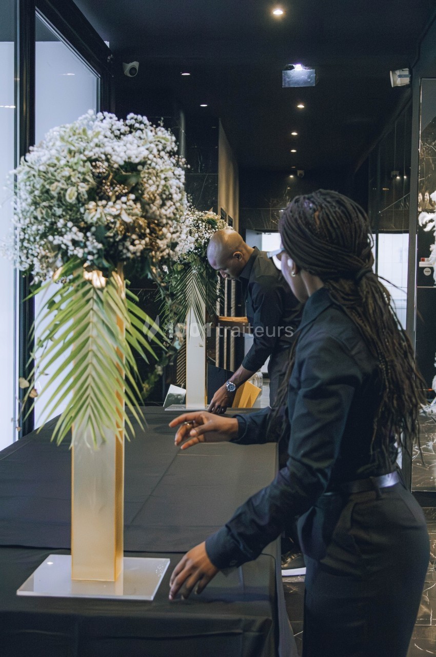 Deux personnes en tenue noire arrangent une table décorée de grands bouquets floraux dans un espace intérieur sombre.