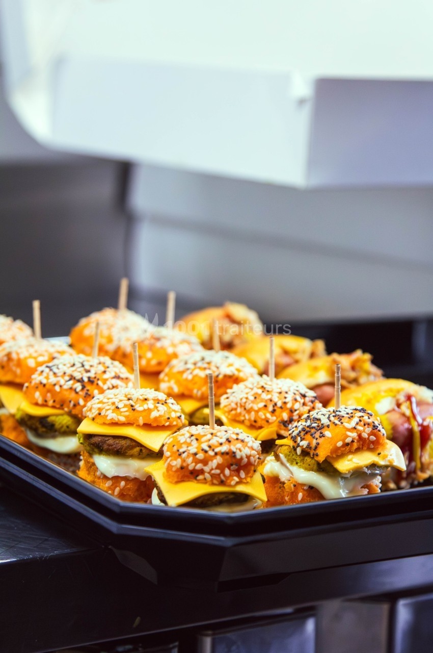 Plateau de mini-burgers garnis de fromage et de graines de sésame, présentés sur un plan de travail en cuisine.