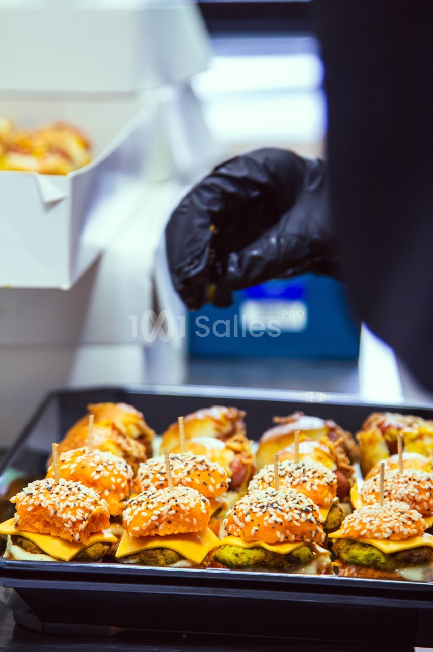 Plateau de mini-burgers garnis de fromage et de graines de sésame, préparés par une main gantée en cuisine.