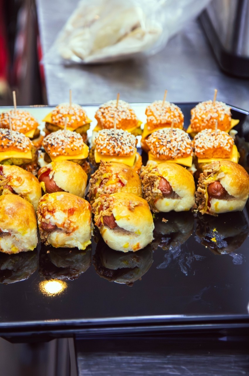 Plateau de mini-sandwichs variés, garnis de fromage, viande hachée et saucisses, présentés sur une table.