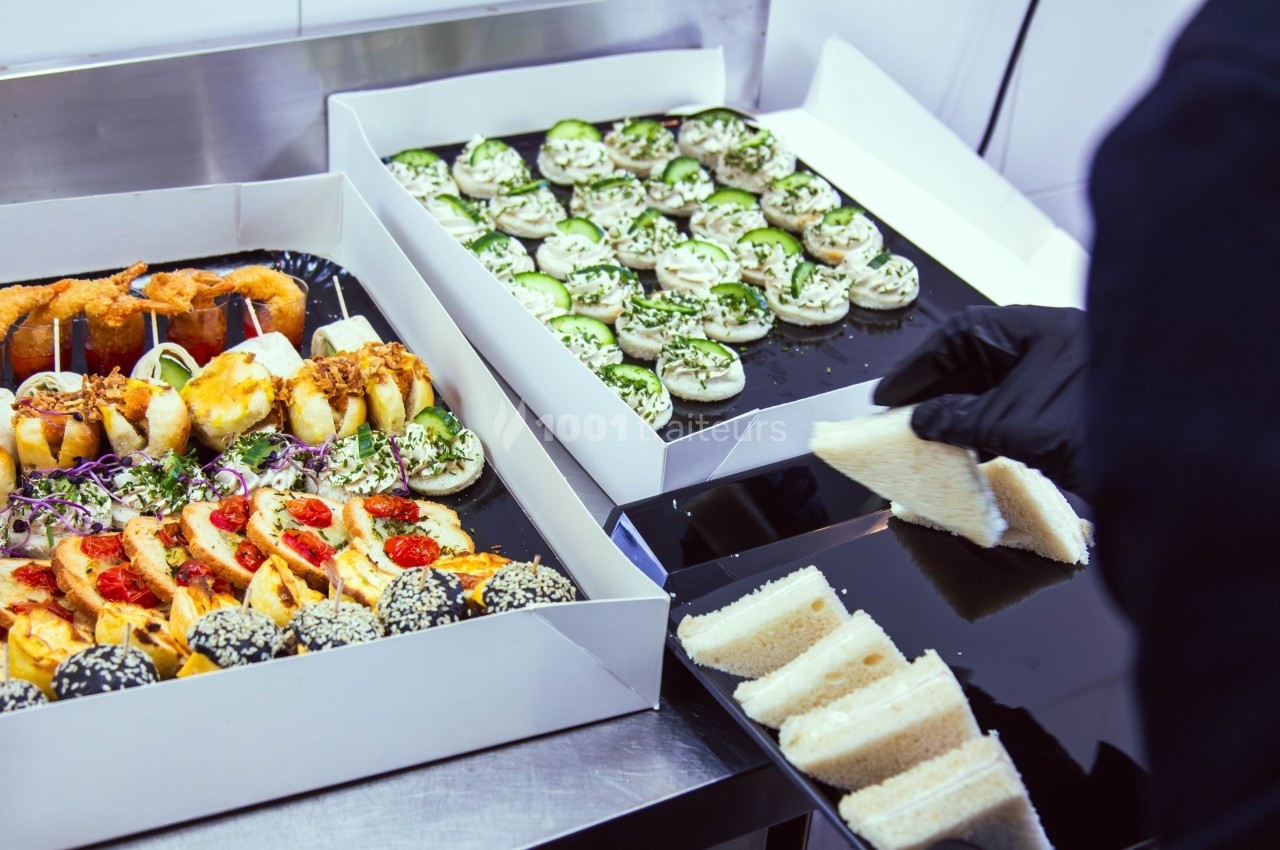 Plateaux de bouchées variées et sandwichs préparés dans une cuisine, une personne en gants noirs les dispose.