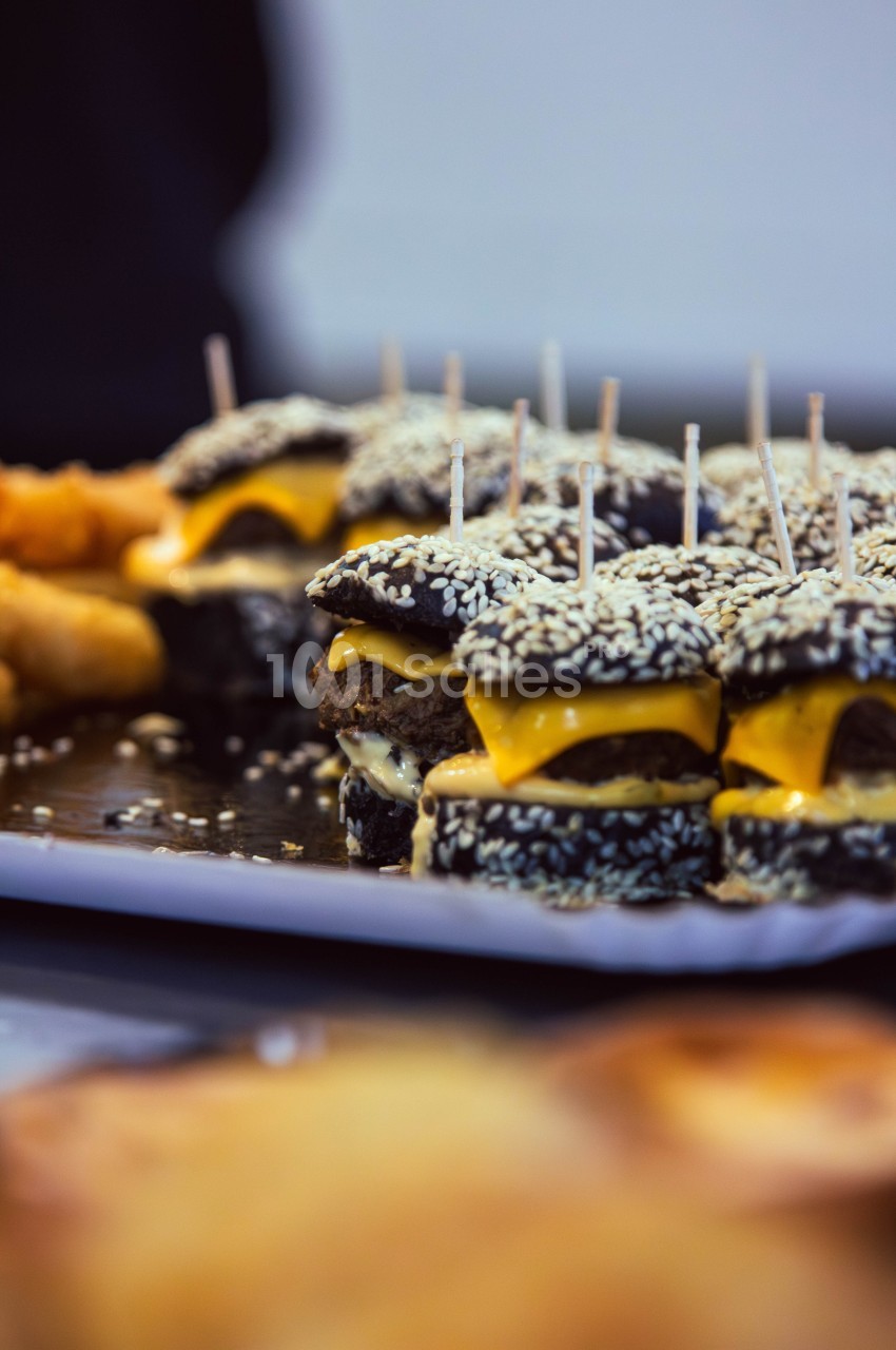 Mini-burgers au pain noir et graines de sésame, garnis de fromage et servis sur un plateau avec des amuse-bouches.