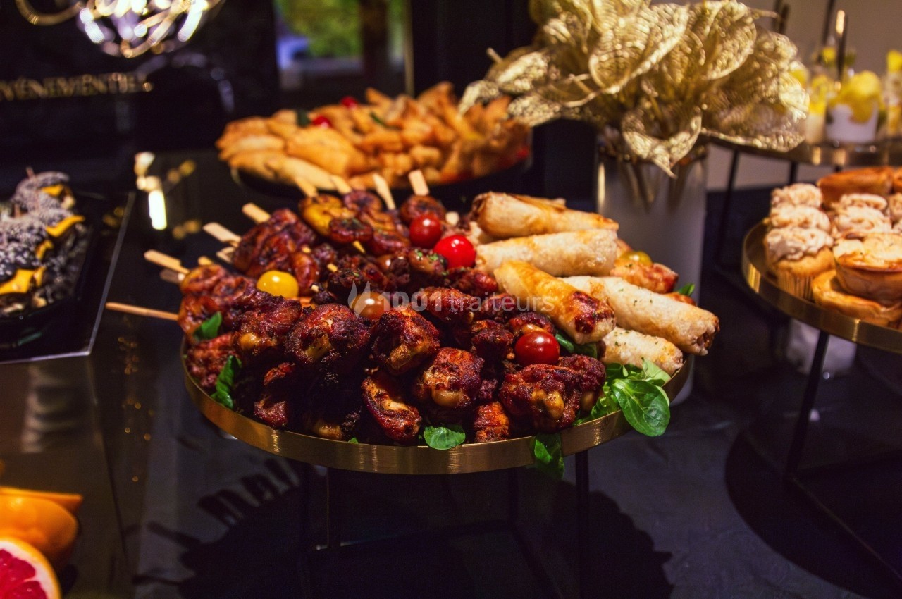 Plateau de buffet avec brochettes de viande, rouleaux frits, et décorations dorées sur une table sombre.