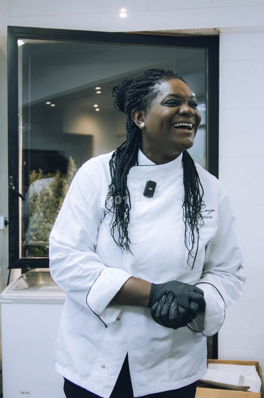 Une cheffe en veste blanche sourit dans une cuisine moderne, portant des gants noirs et un micro-cravate.