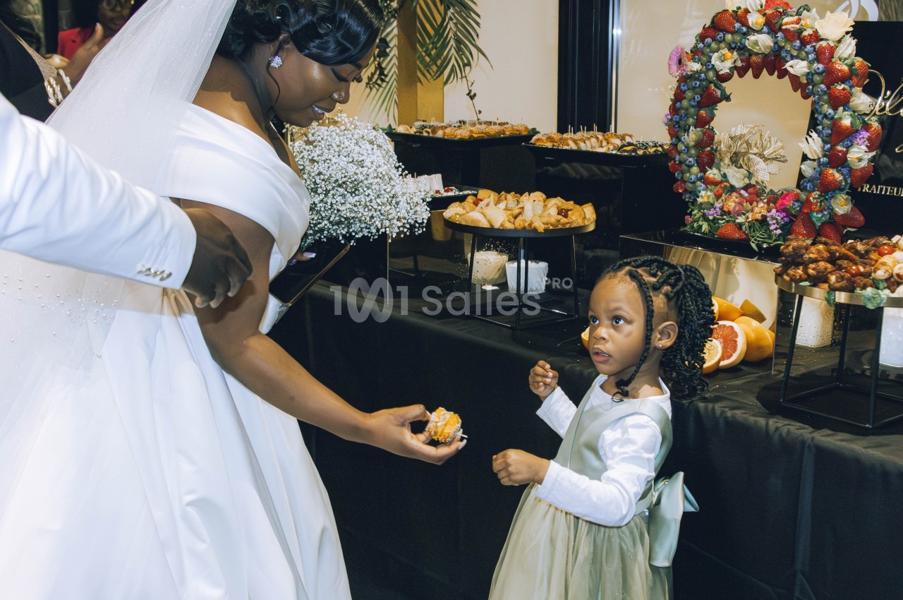 Une mariée tend un dessert à une petite fille en robe verte devant une table garnie de pâtisseries.