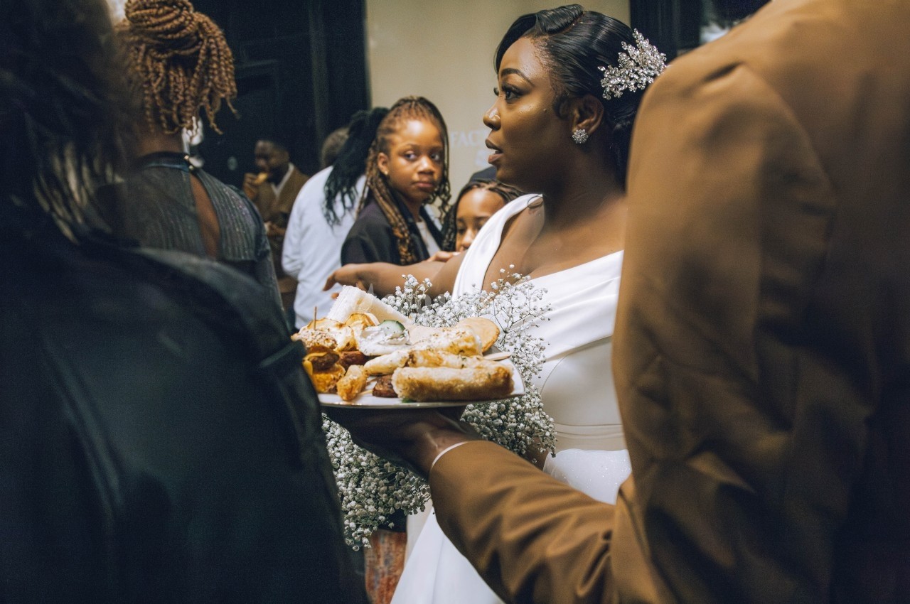 Une femme en robe blanche tient un plateau de pâtisseries entourée de personnes lors d'un rassemblement.
