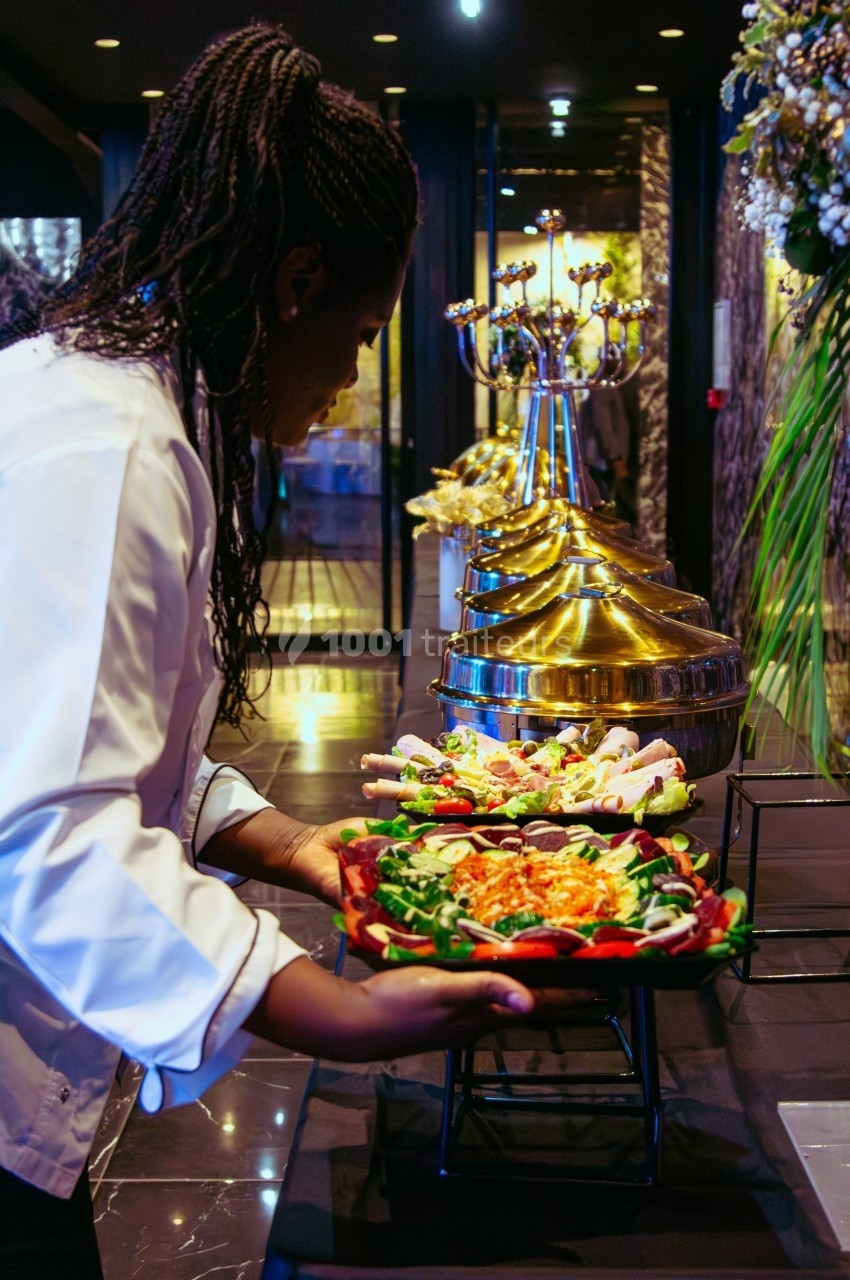 Une cheffe dispose un plat de salade sur une table de buffet garnie de mets variés dans un cadre élégant.