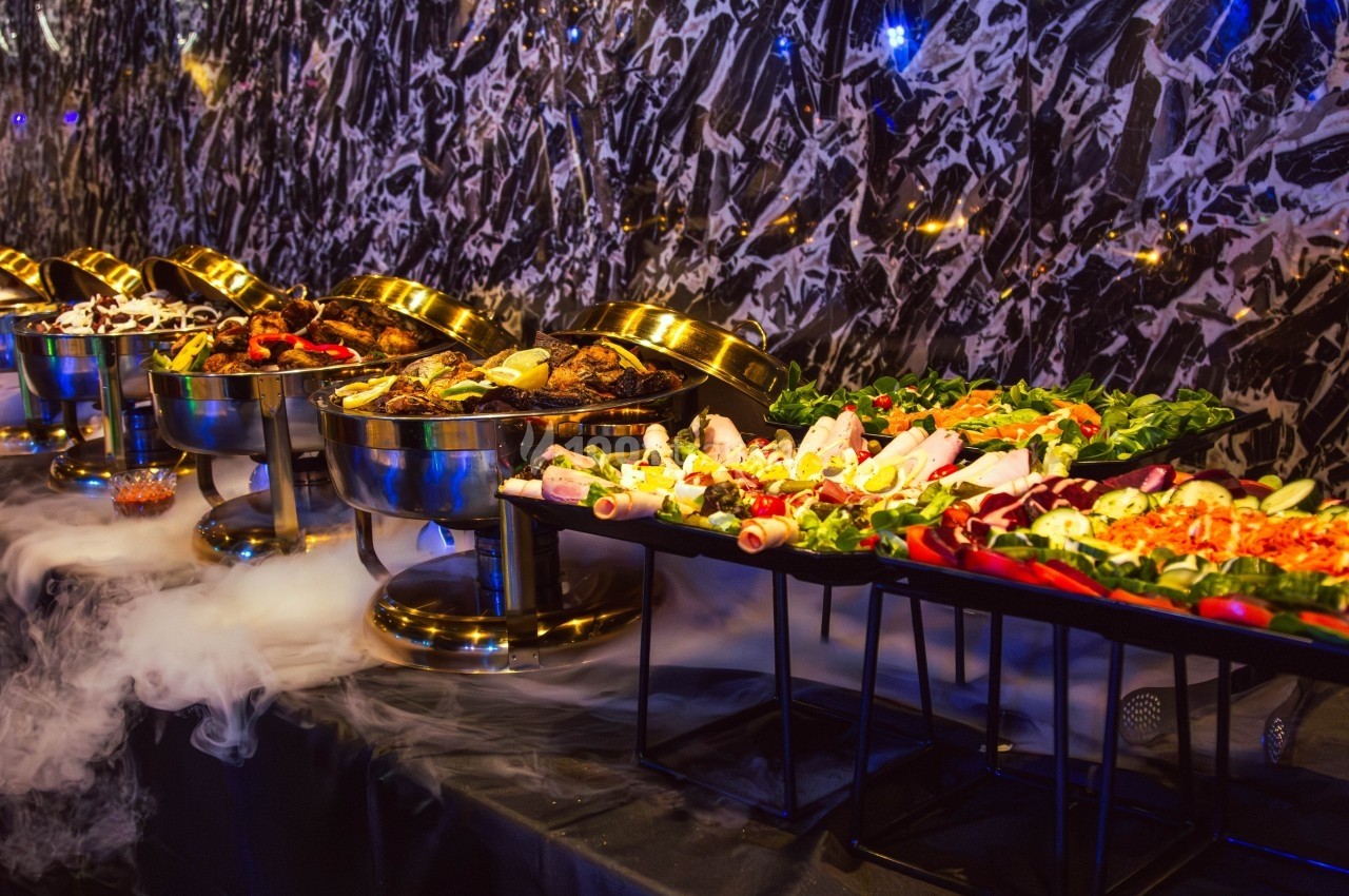 Buffet varié avec plats chauds dans des récipients en métal et salades fraîches sur des plateaux, décoré de fumée.