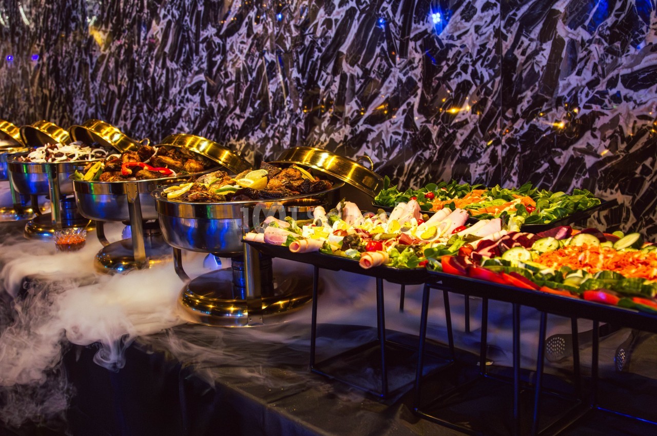 Buffet varié avec plats chauds dans des récipients en métal et salades fraîches sur des plateaux, décoré de fumée.
