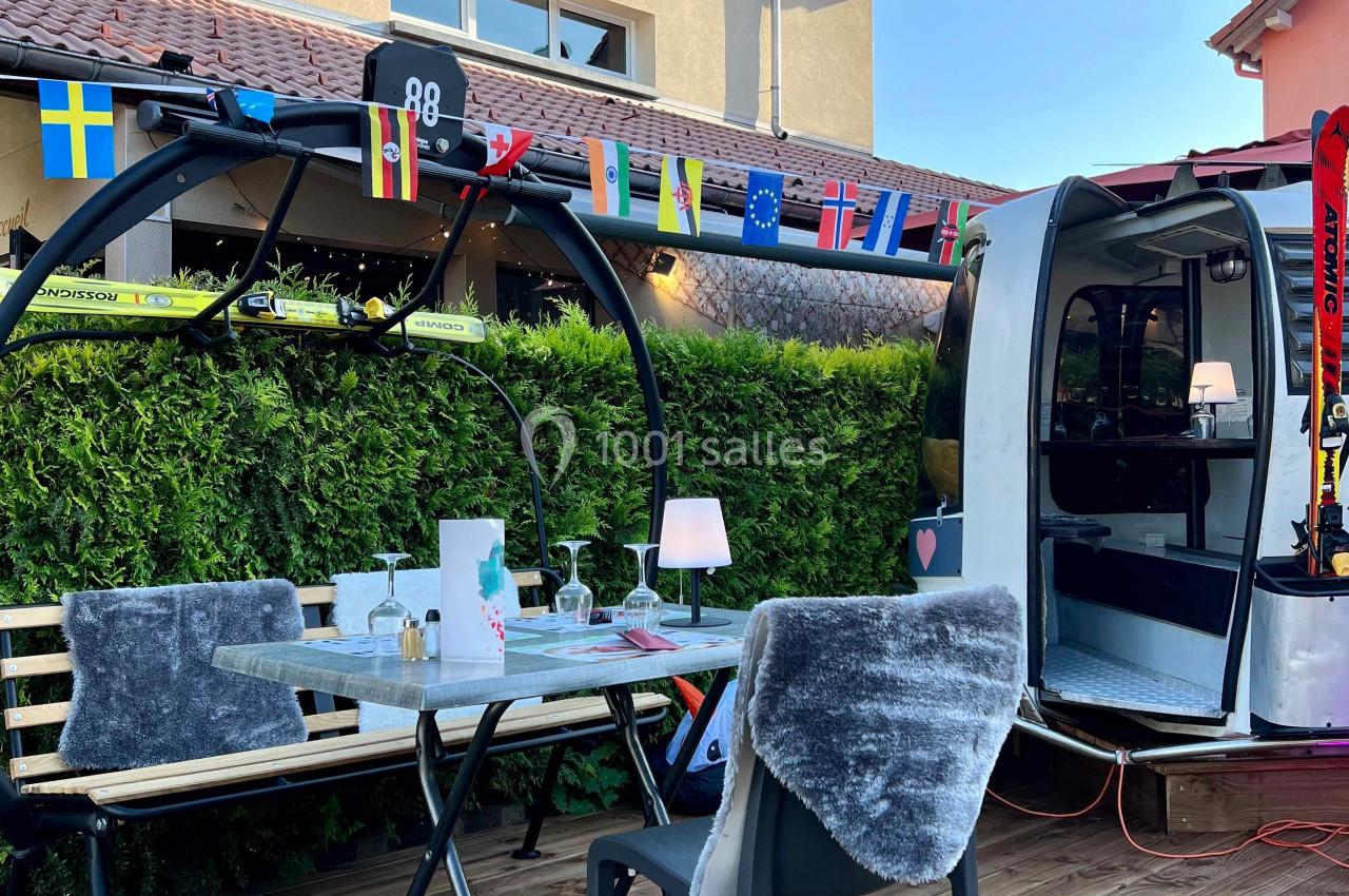 Terrasse en bois avec table dressée, chaises, télécabine aménagée et guirlande de drapeaux devant un bâtiment.