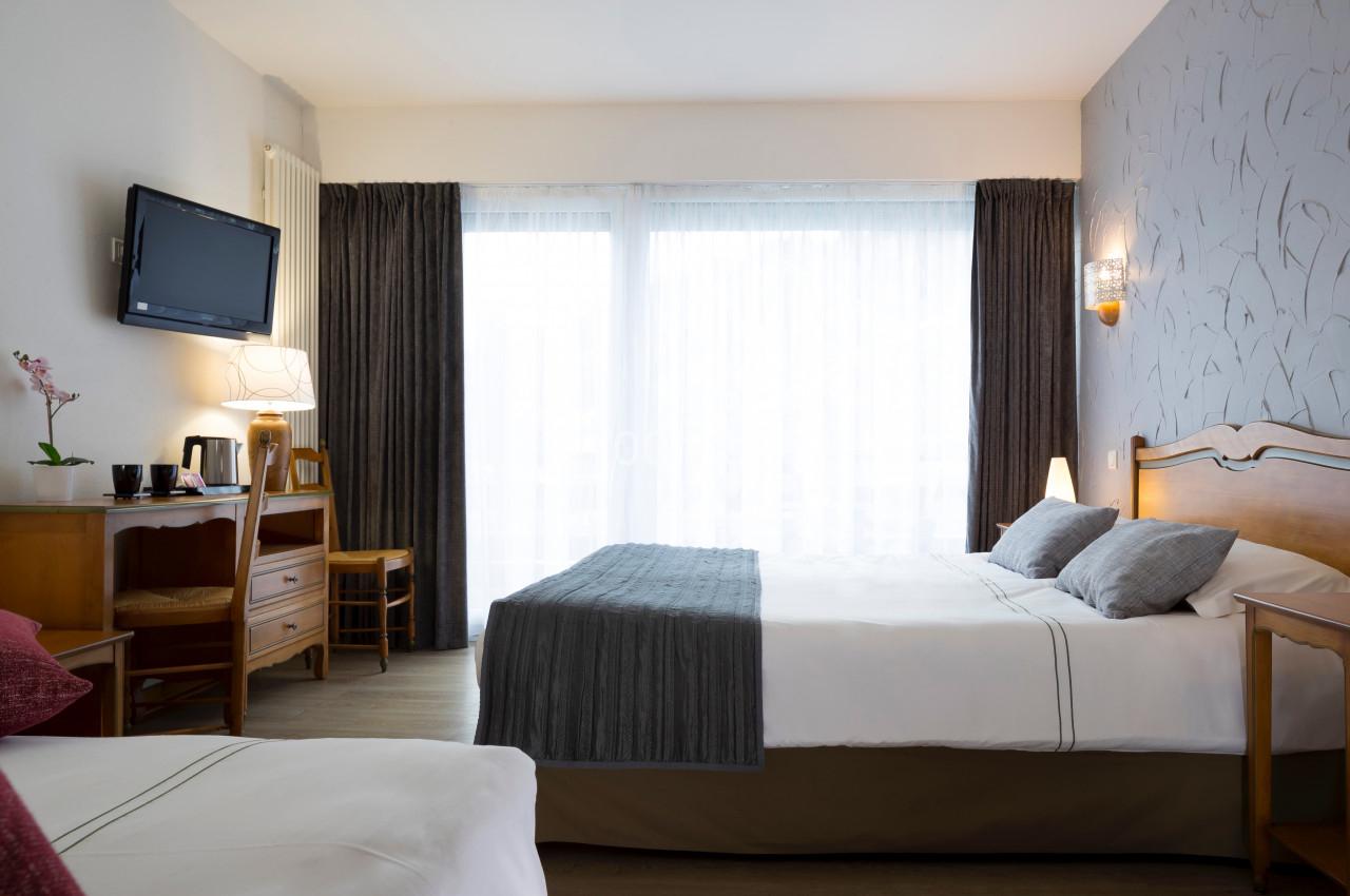 Chambre d'hôtel lumineuse avec deux lits, bureau en bois, télévision murale et grande fenêtre avec rideaux.