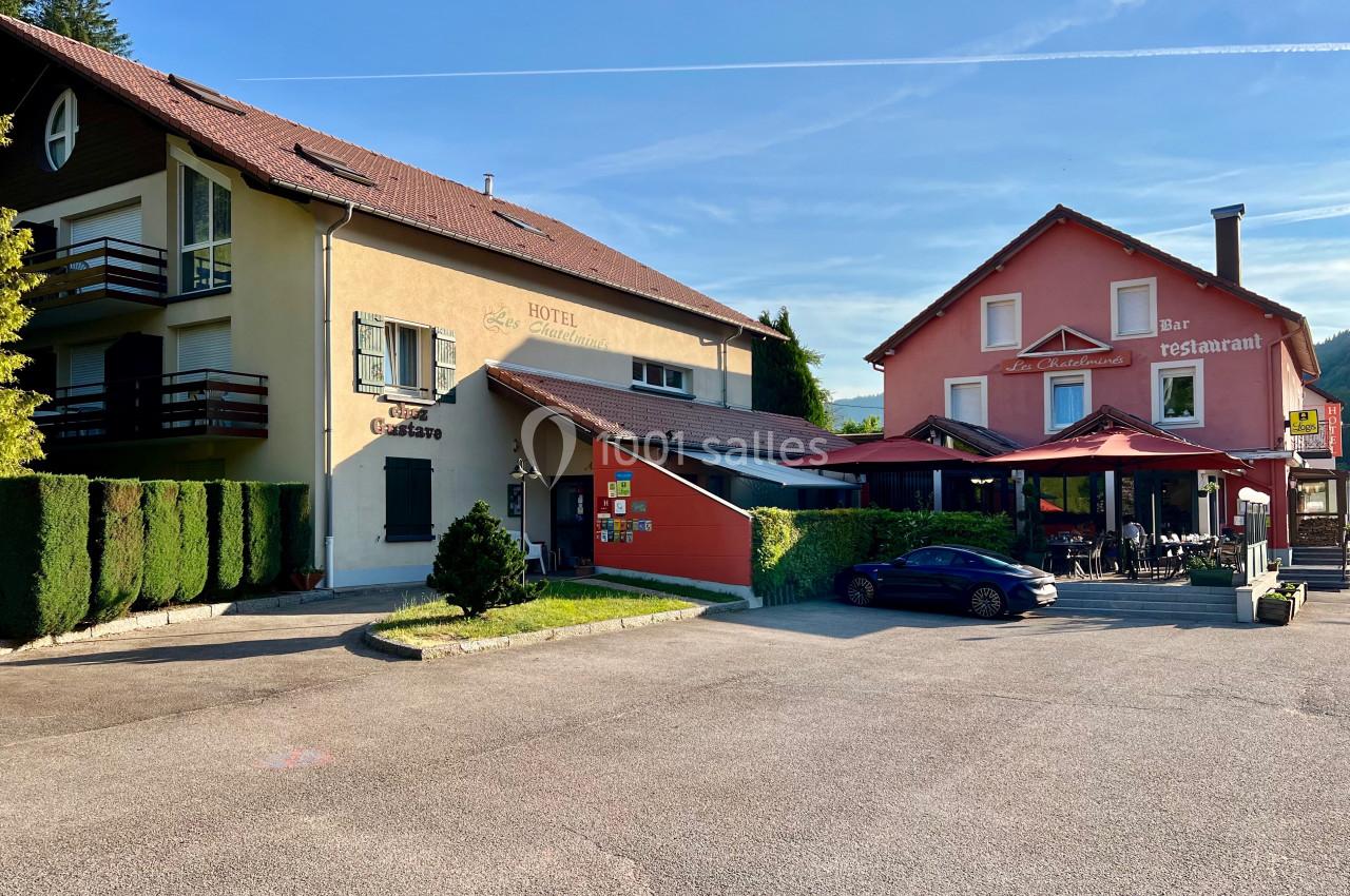 Façade d'un hôtel-restaurant avec terrasse, entouré de végétation et d'un parking sous un ciel dégagé.